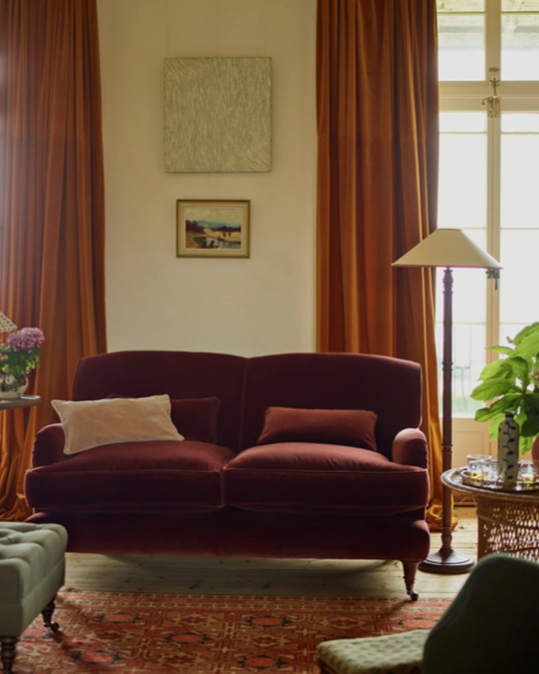 Living room scene with a plush two-seater maroon couch, decorative pillows, and artistic wall decor in a warm, inviting light.