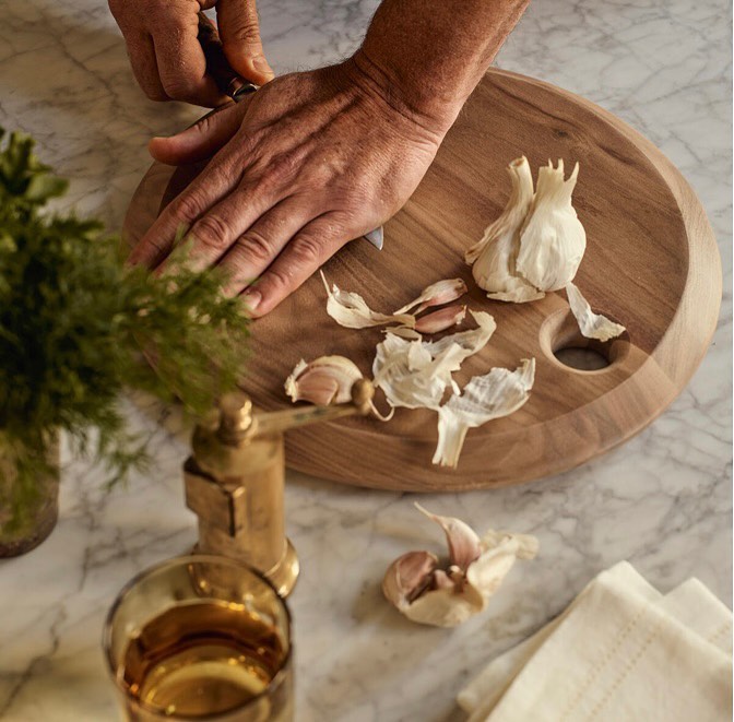Hands prepare garlic on a wooden cutting board with knife on a marble surface, with greenery and drinking glass in the background.