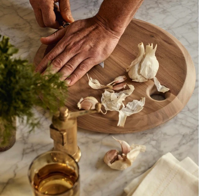 Hands prepare garlic on a wooden cutting board with knife on a marble surface, with greenery and drinking glass in the background.