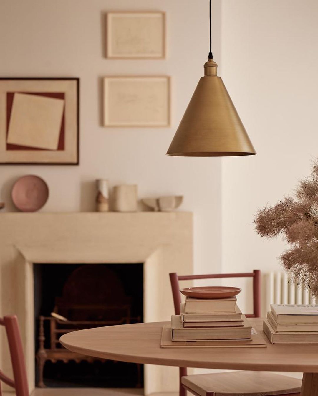 Interior photo of a dining room showcasing a gold pendant light, round wooden table with books, and a fireplace in a neutral color palette.