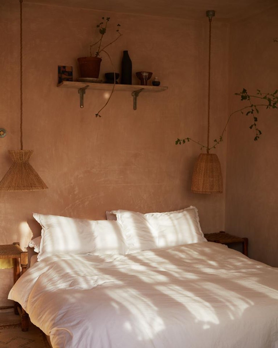 Bedroom interior with a white bed bathed in natural light, rattan pendant lights, and a shelf with decorative items in a warm-toned room.