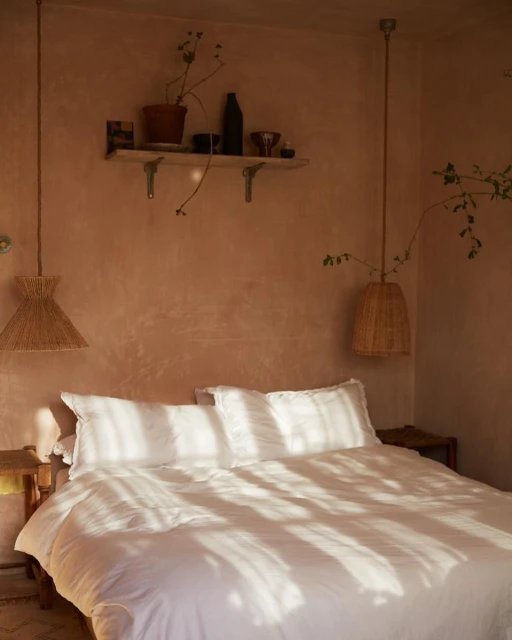 Bedroom interior with a white bed bathed in natural light, rattan pendant lights, and a shelf with decorative items in a warm-toned room.
