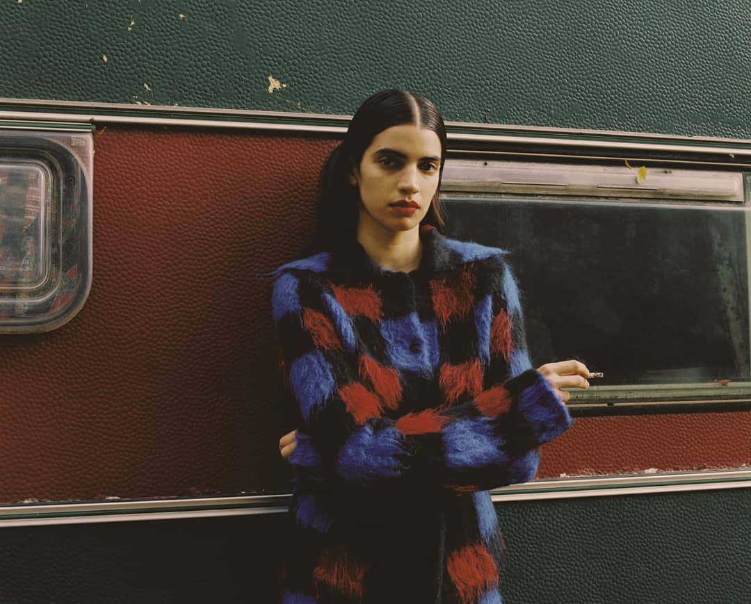 A young woman with dark hair and red lipstick poses in front of a green and maroon trailer, wearing a blue, red, and black fuzzy jacket.