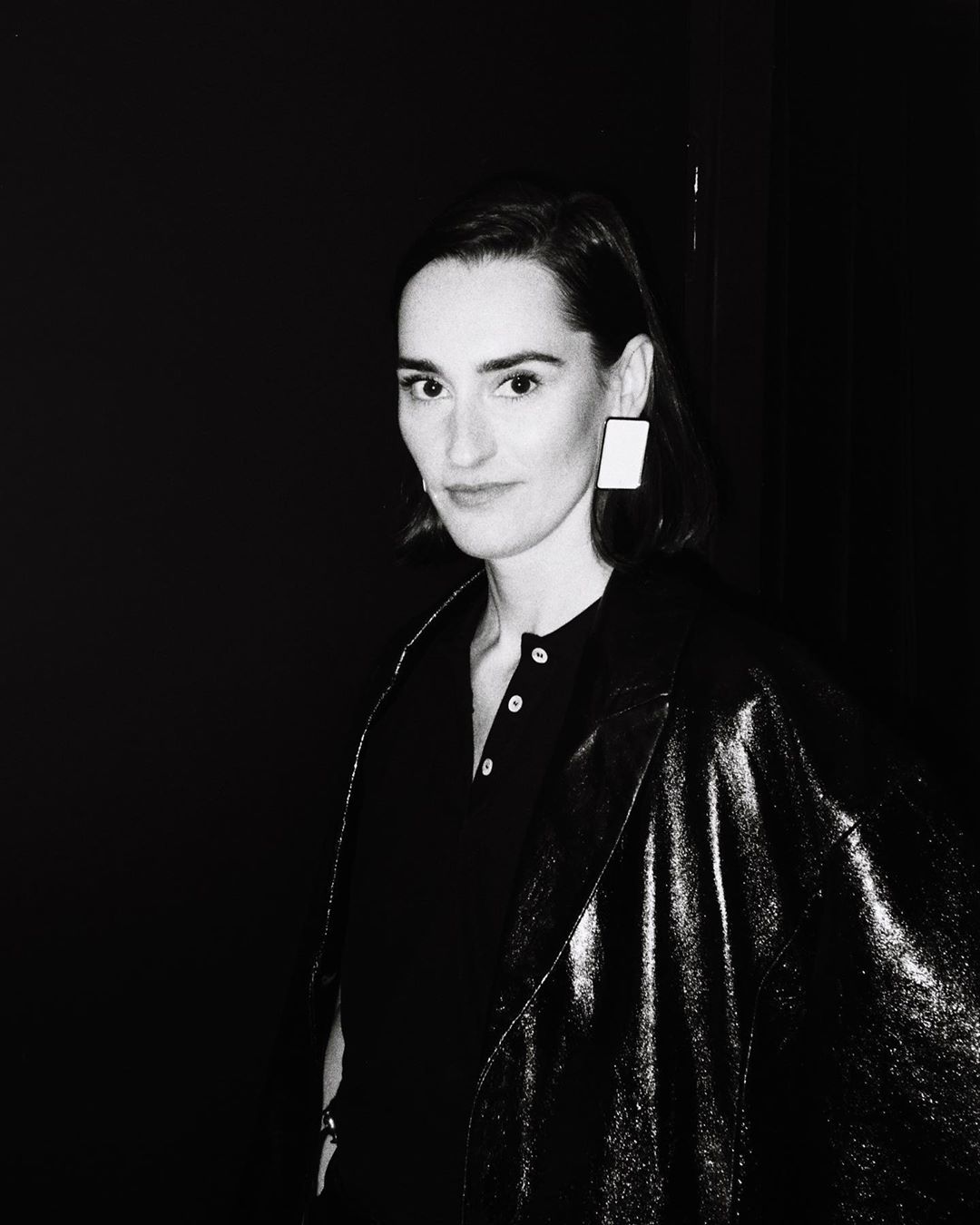 Portrait of an adult female in a dark jacket and square earrings against a black backdrop in a black and white photograph.