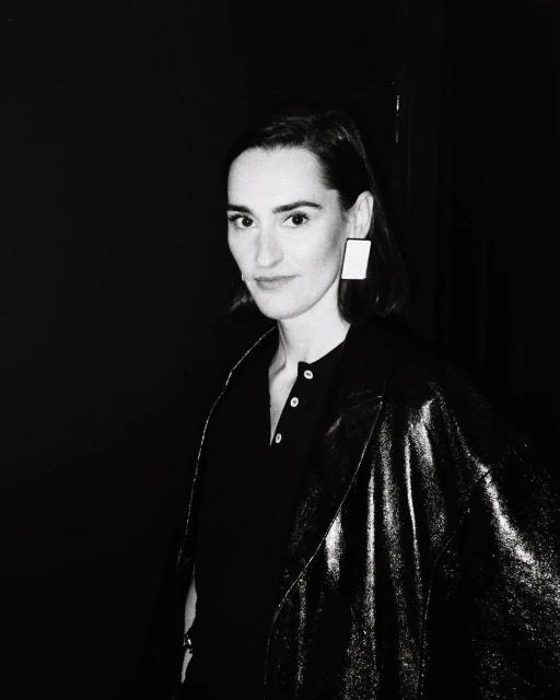 Portrait of an adult female in a dark jacket and square earrings against a black backdrop in a black and white photograph.