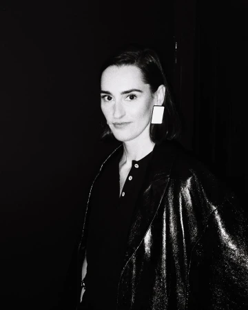 Portrait of an adult female in a dark jacket and square earrings against a black backdrop in a black and white photograph.