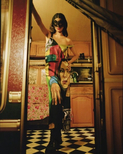 Woman in printed dress and lace mask posing indoors. Checkerboard floors, warm lighting, and retro interior design details visible.