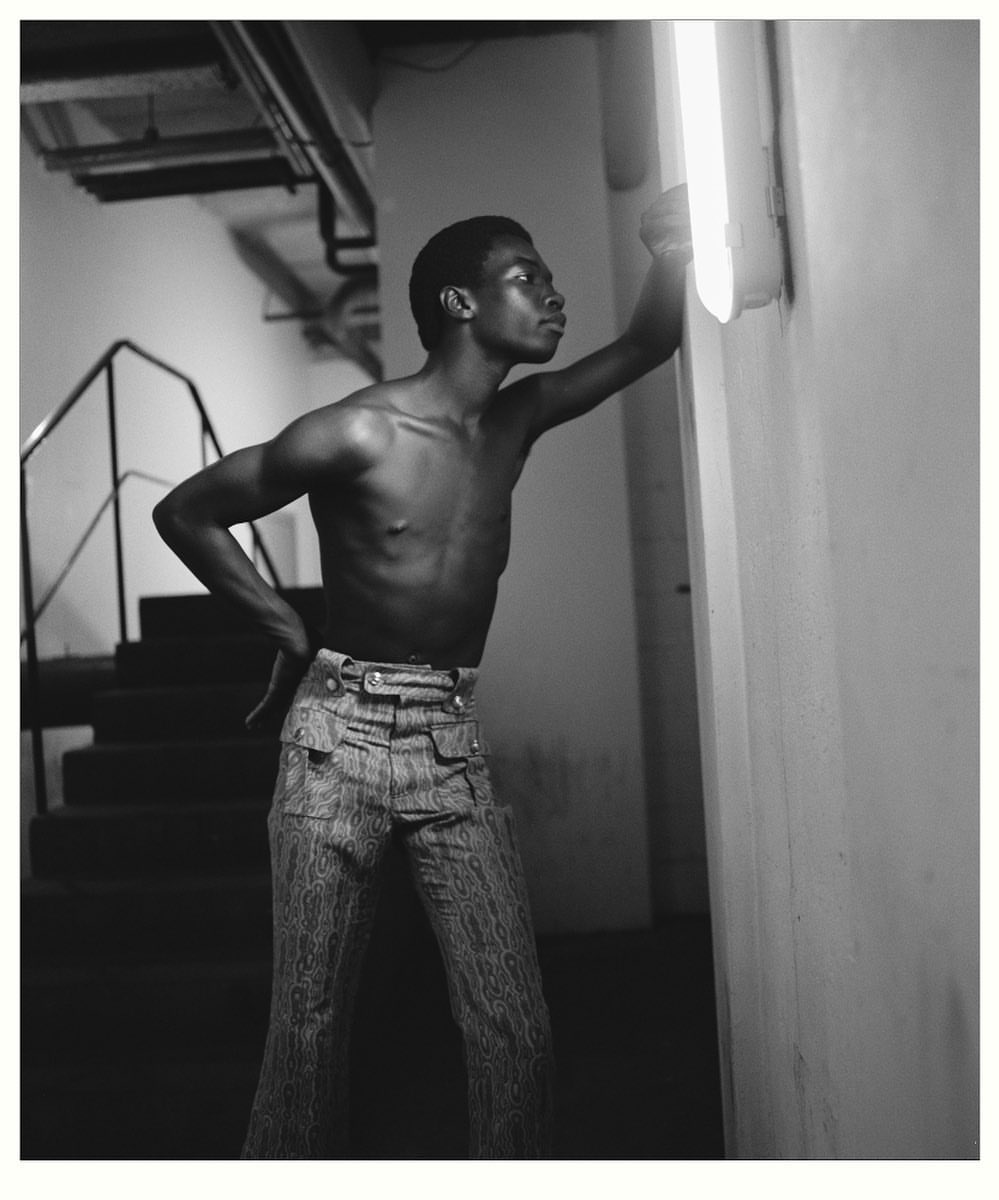Black and white portrait of a shirtless man in patterned pants, leaning against a white wall with an overhead fluorescent light.