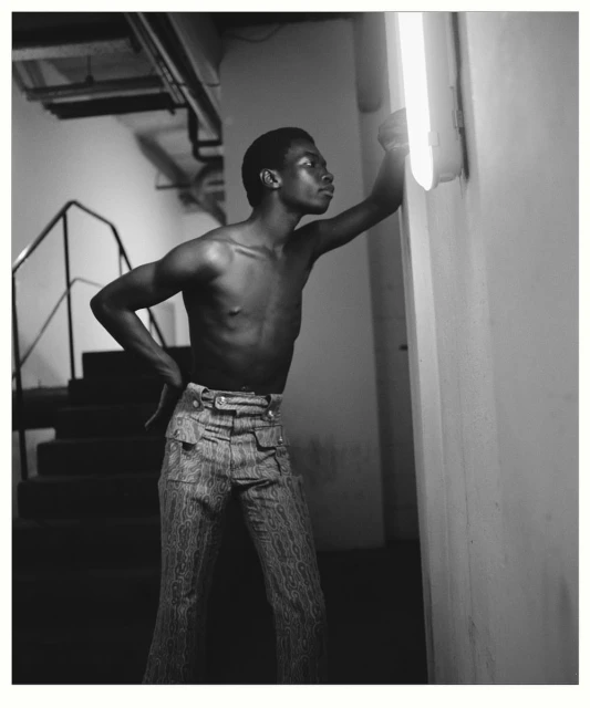 Black and white portrait of a shirtless man in patterned pants, leaning against a white wall with an overhead fluorescent light.