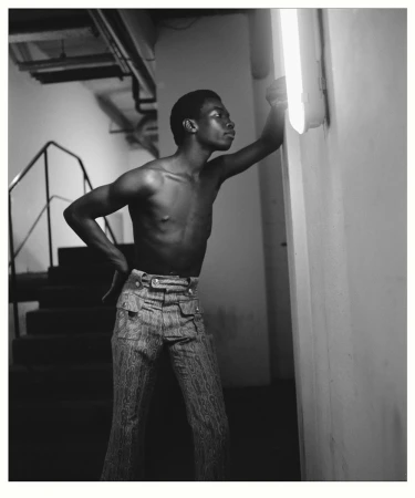 Black and white portrait of a shirtless man in patterned pants, leaning against a white wall with an overhead fluorescent light.
