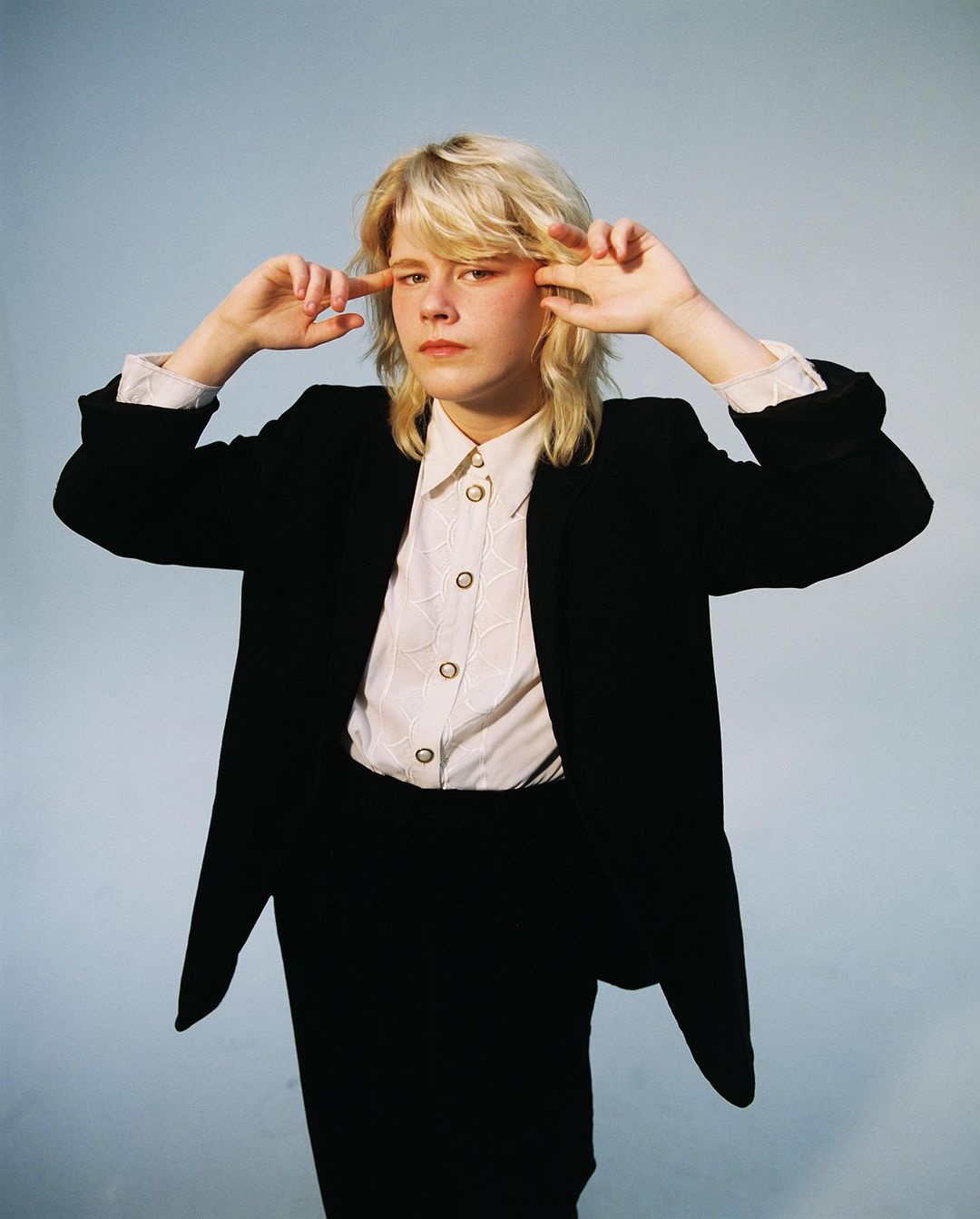 Blonde person wearing a black blazer and white button shirt pointing at temples against a light blue backdrop.