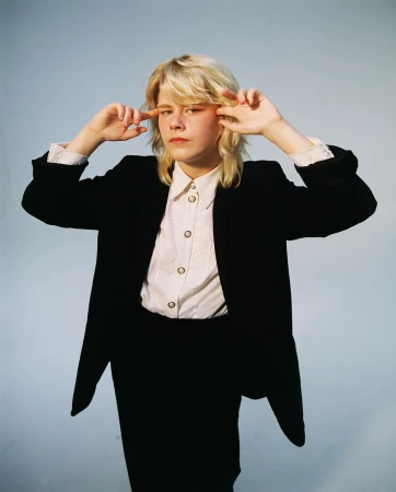 Blonde person wearing a black blazer and white button shirt pointing at temples against a light blue backdrop.