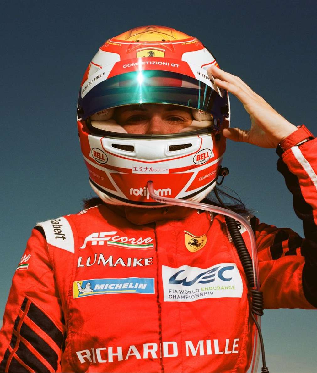 Close-up of a person in a red racing suit and helmet against a blue sky, hand raised near the helmet, looking directly at the viewer.