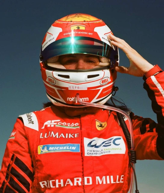 Close-up of a person in a red racing suit and helmet against a blue sky, hand raised near the helmet, looking directly at the viewer.