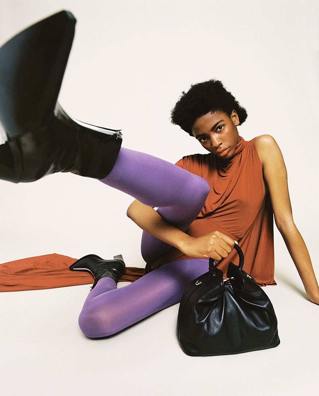 A woman wearing a brown top and violet leggings poses on a white surface, holding a black handbag with a single black boot in the foreground.