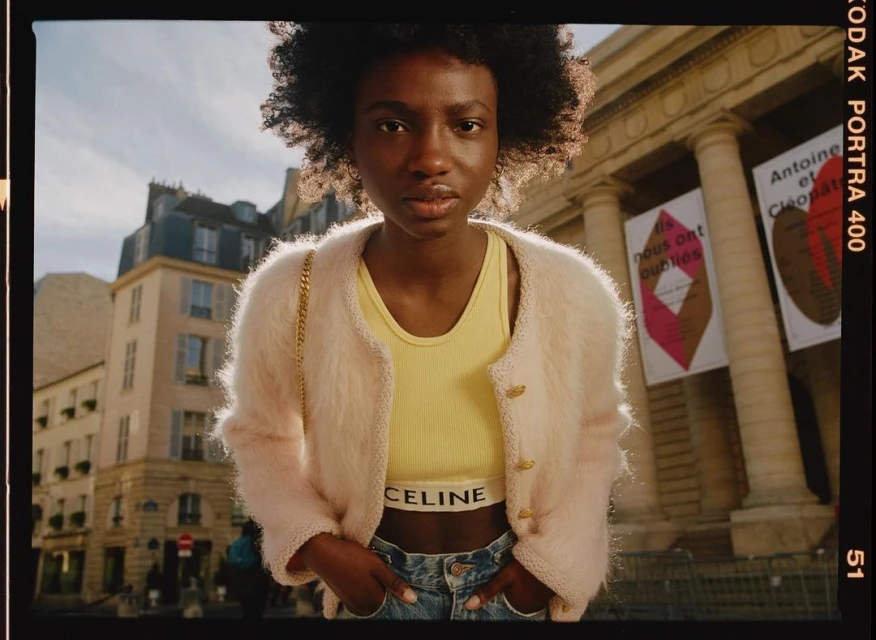 Young Black woman in stylish outfit poses outdoors in front of classical building with posters. Soft natural light. Fashion editorial shot.