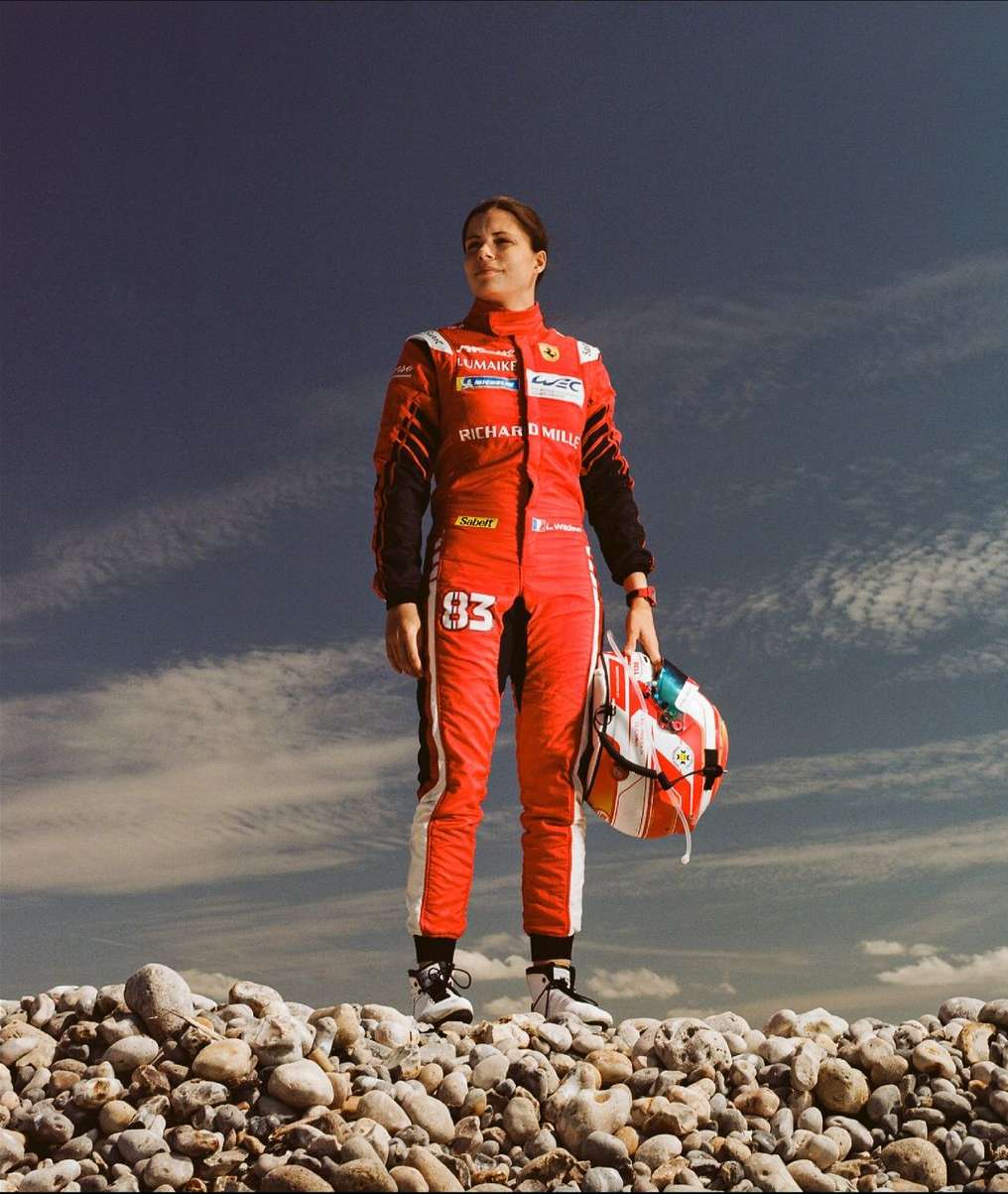 A female race car driver in a red suit holds her helmet while standing on a rocky surface against a partly cloudy blue sky.