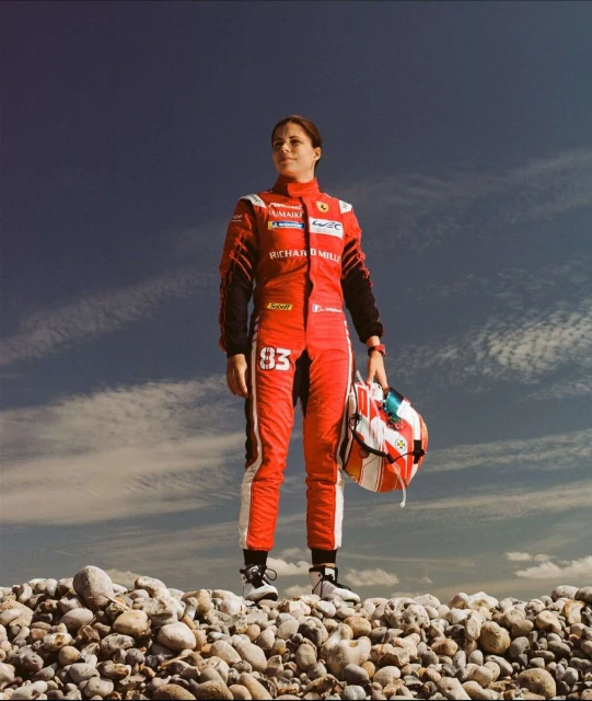 A female race car driver in a red suit holds her helmet while standing on a rocky surface against a partly cloudy blue sky.