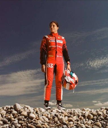 A female race car driver in a red suit holds her helmet while standing on a rocky surface against a partly cloudy blue sky.