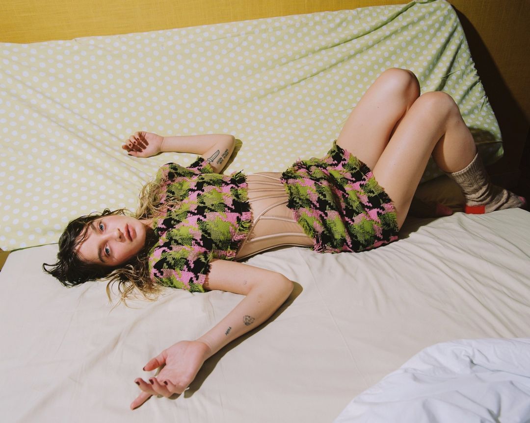 A young woman with fair skin lies on her back on a bed with a patterned comforter, wearing a knit co-ord set and socks.