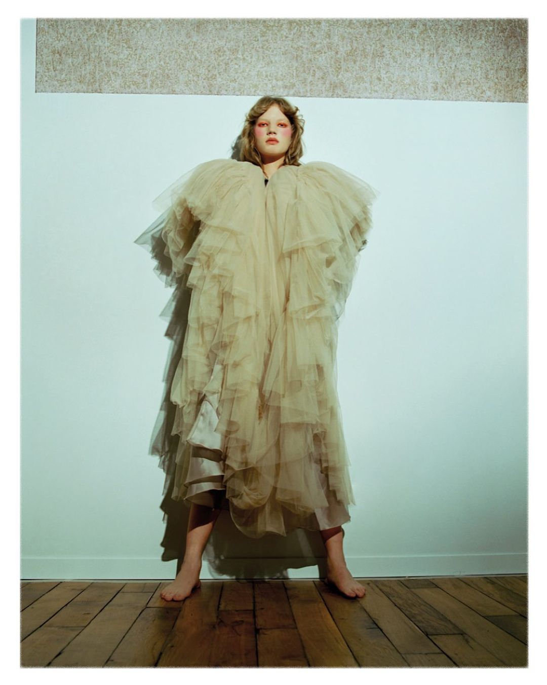 Woman in a layered beige tulle gown poses barefoot in a studio with hardwood floors and a cream and grey textured background panel.