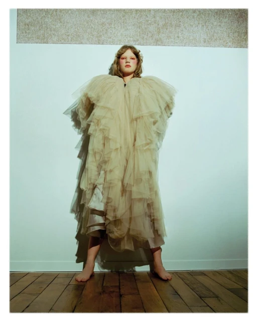 Woman in a layered beige tulle gown poses barefoot in a studio with hardwood floors and a cream and grey textured background panel.