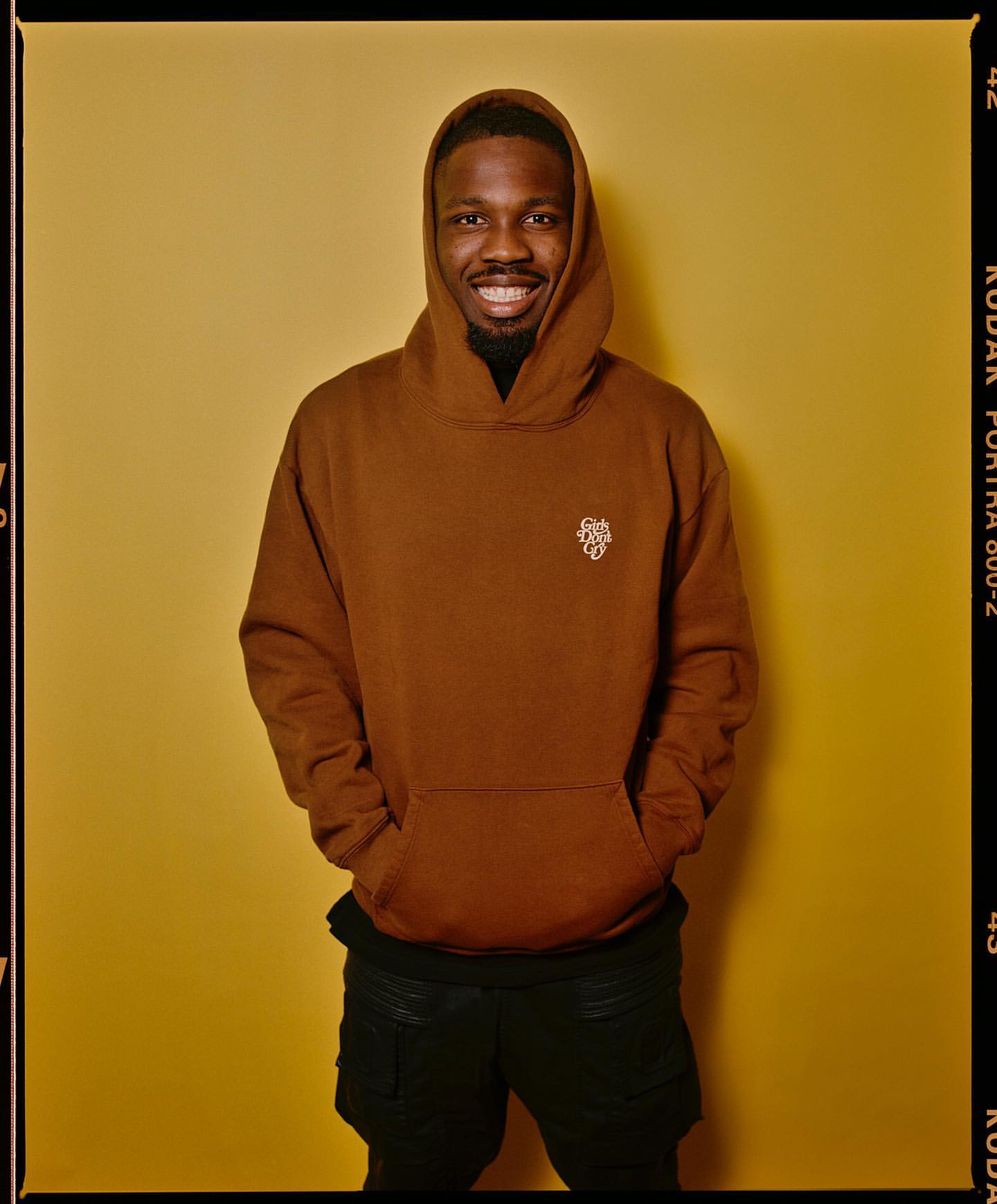 Portrait of a smiling man wearing a brown hooded sweatshirt in front of a tan-colored wall with a dark-colored film border.