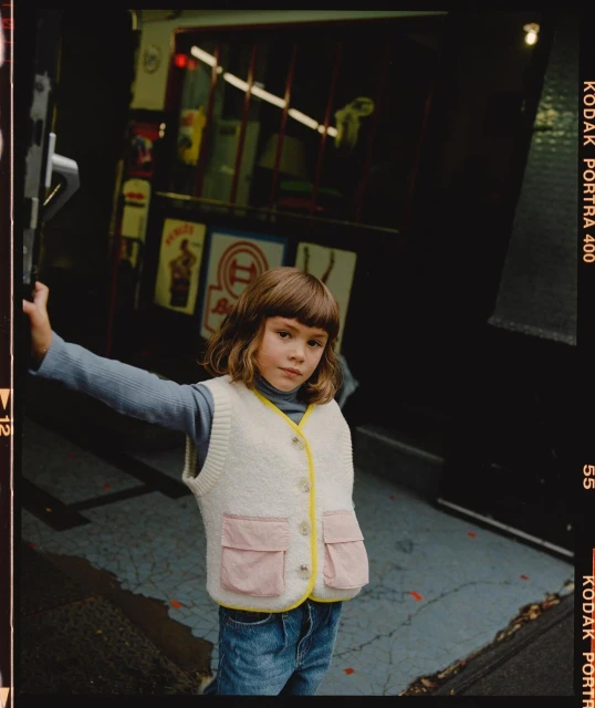 A child with bangs wearing a cream-colored vest stands in front of a dark building with posters, a Kodak Portra 400 film border.