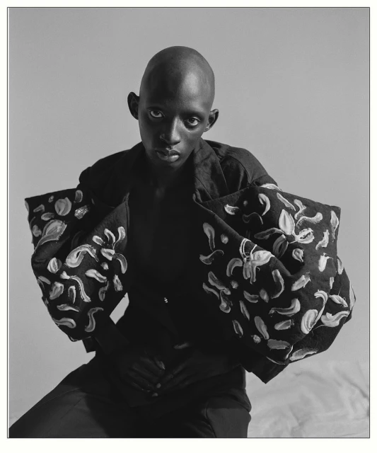 Striking black-and-white portrait of a bald Black adult in a fashion-forward floral accented jacket looking directly at the camera indoors.