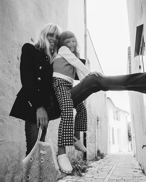 Black and white shot of adult female in black jacket with child wearing checkered pants sitting on wall in alleyway with crochet bag.