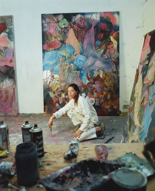 Female artist in white overalls crouches on floor with art supplies and paint cans, working on large abstract paintings in her studio.
