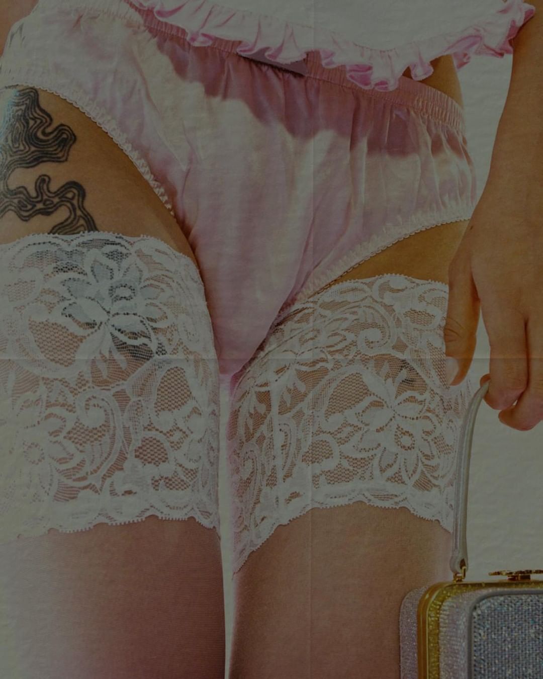 Woman posing in lingerie with floral lace stockings and glittery purse against a solid backdrop, soft lighting, close-up shot.