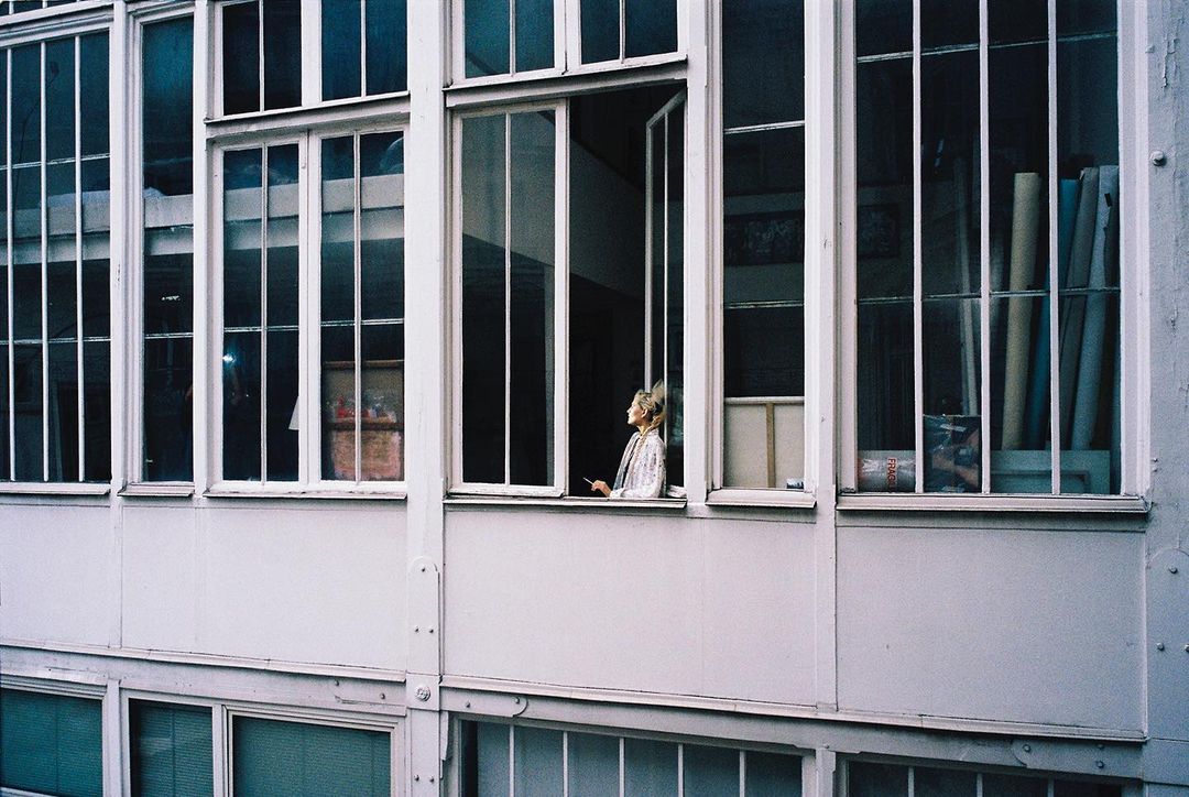 A young adult woman stands in an open window, framed by white window panes, looking out on a sunny day.