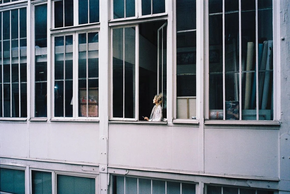 A young adult woman stands in an open window, framed by white window panes, looking out on a sunny day.