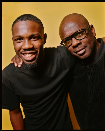 Portrait of two Black men posing closely together against a warm yellow studio backdrop by photographer Emma Picq