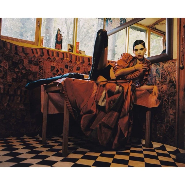 A person in an orange shirt and black boots lies on a table in a room with black and white checkered flooring and patterned walls.