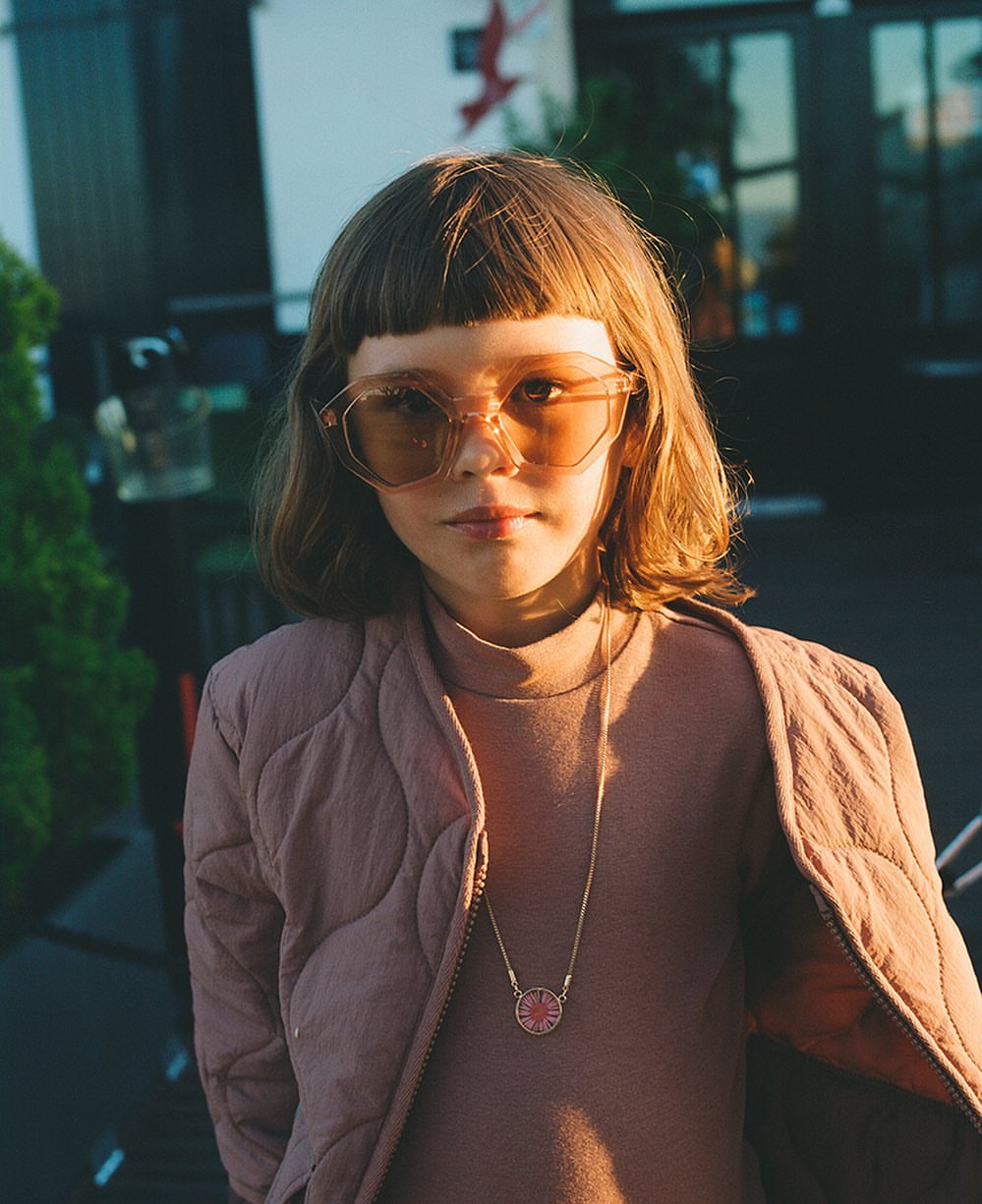 Child with retro sunglasses wearing a quilted jacket outside at dusk with buildings and trees in the background.