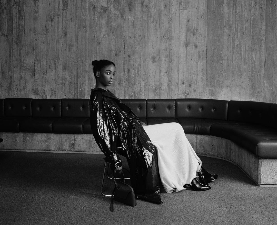 A young woman in a black coat sits in front of a concrete wall, holding a black purse, with a black leather couch in the background.