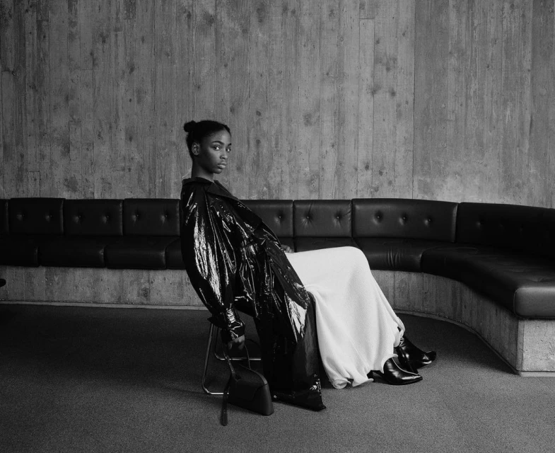 A young woman in a black coat sits in front of a concrete wall, holding a black purse, with a black leather couch in the background.