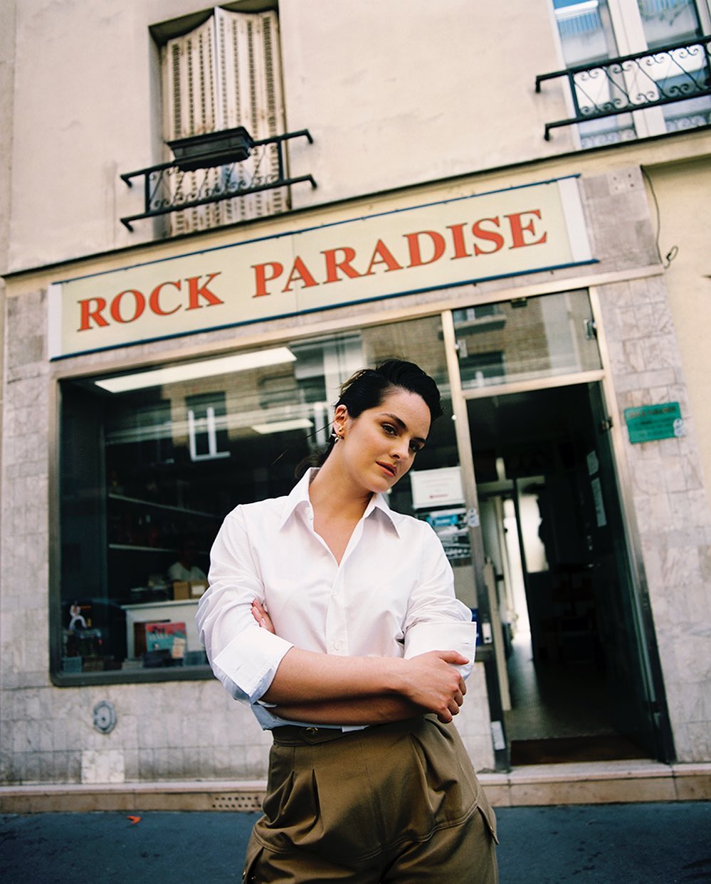 Portrait of a brunette woman with short hair posing in front of a storefront with a white shirt and khaki pants in an urban setting.