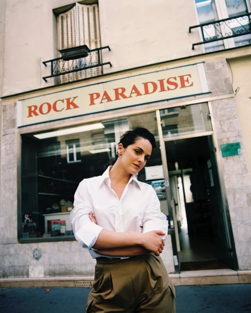 Portrait of a brunette woman with short hair posing in front of a storefront with a white shirt and khaki pants in an urban setting.
