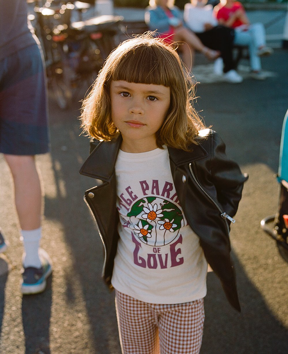 Young girl in a leather jacket stands outside wearing a graphic t-shirt and patterned pants, lit by sunlight. Pedestrians in background.
