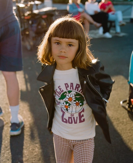 Young girl in a leather jacket stands outside wearing a graphic t-shirt and patterned pants, lit by sunlight. Pedestrians in background.