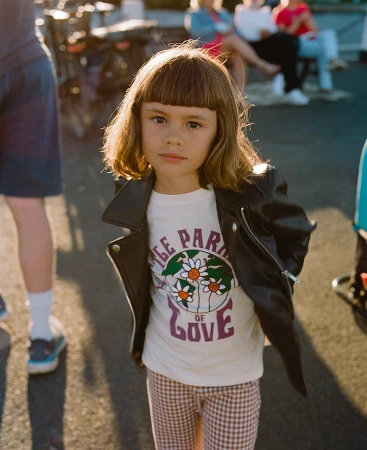 Young girl in a leather jacket stands outside wearing a graphic t-shirt and patterned pants, lit by sunlight. Pedestrians in background.