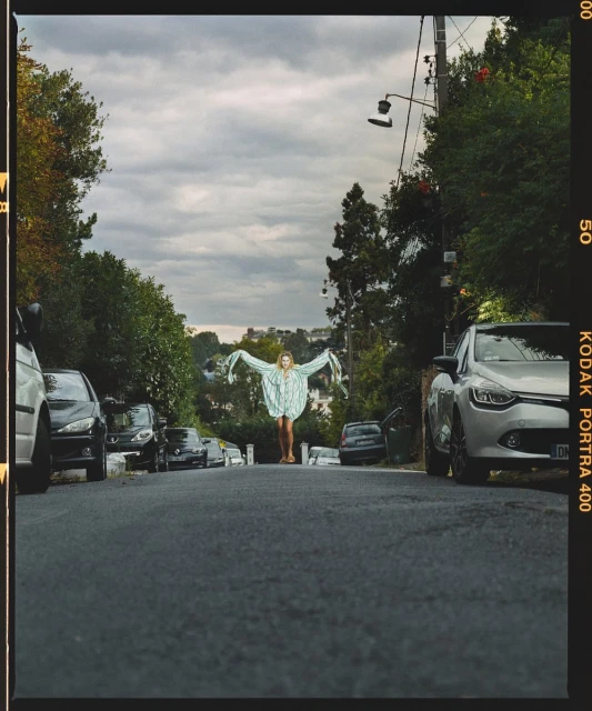 Woman in an oversized shirt stands in the center of a residential street, surrounded by parked cars and greenery against an overcast sky.