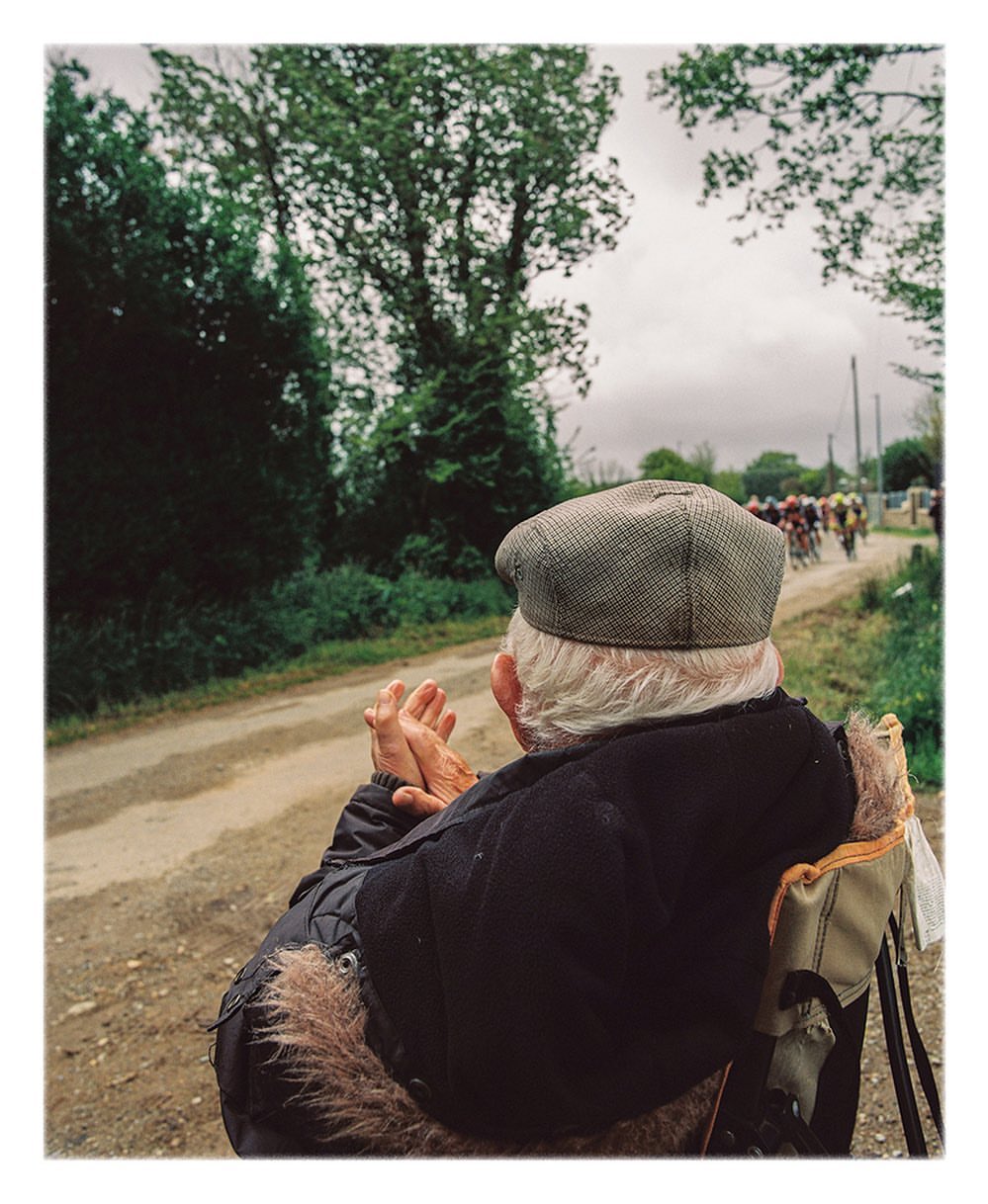 Elderly man in cap watching a bicycle race on a rural road lined with trees and vegetation under an overcast sky.