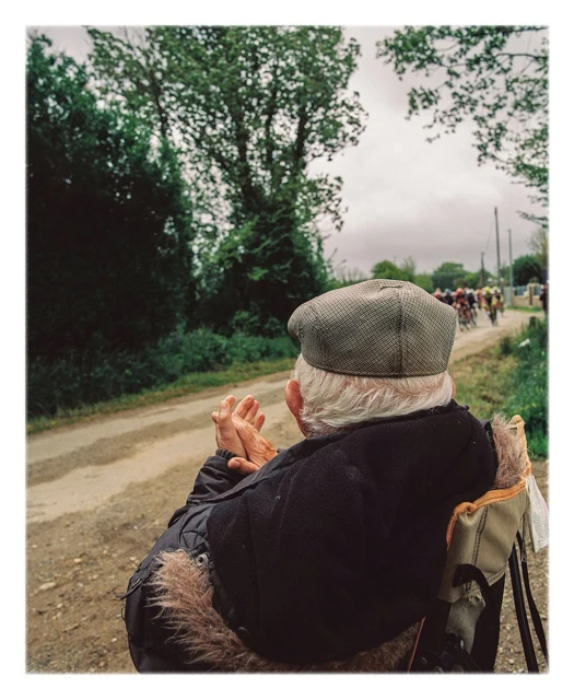Elderly man in cap watching a bicycle race on a rural road lined with trees and vegetation under an overcast sky.