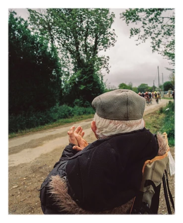 Elderly man in cap watching a bicycle race on a rural road lined with trees and vegetation under an overcast sky.