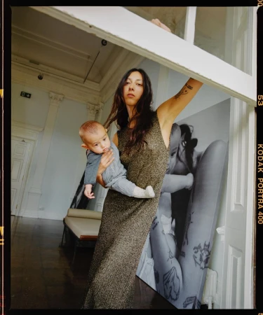 Woman in a shimmering gown stands near a window holding a baby; interior with poster backdrop and natural light streams through.