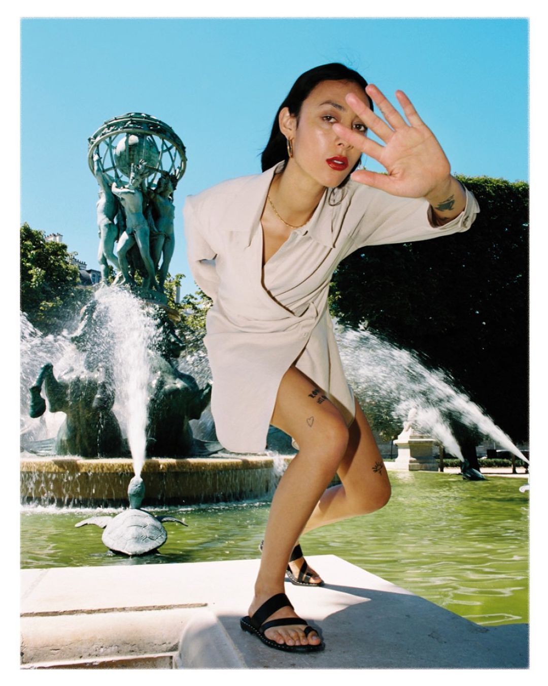 Woman in a short beige dress poses near a fountain and garden on a sunny day, blocking the sun with her hand in an eye-level shot.